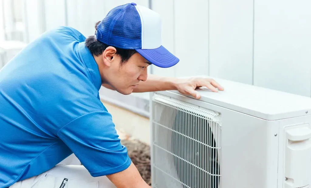 青い作業着とキャップを着たスタッフがエアコン室外機を点検している様子